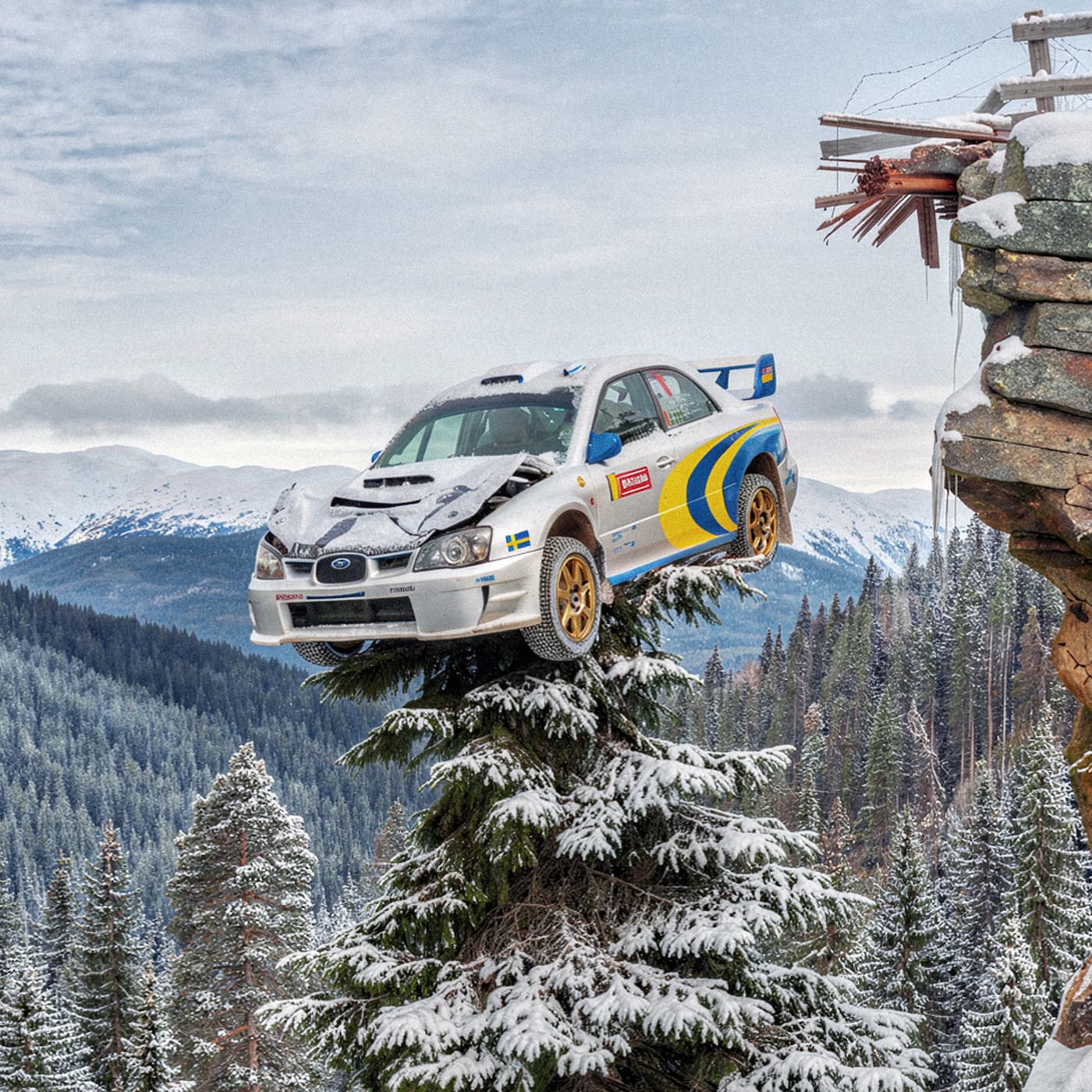 Silverfärgad rallybil med blågula detaljer balanserar på toppen av en snötäckt gran, högt ovanför en skog med snöklädda berg i bakgrunden.